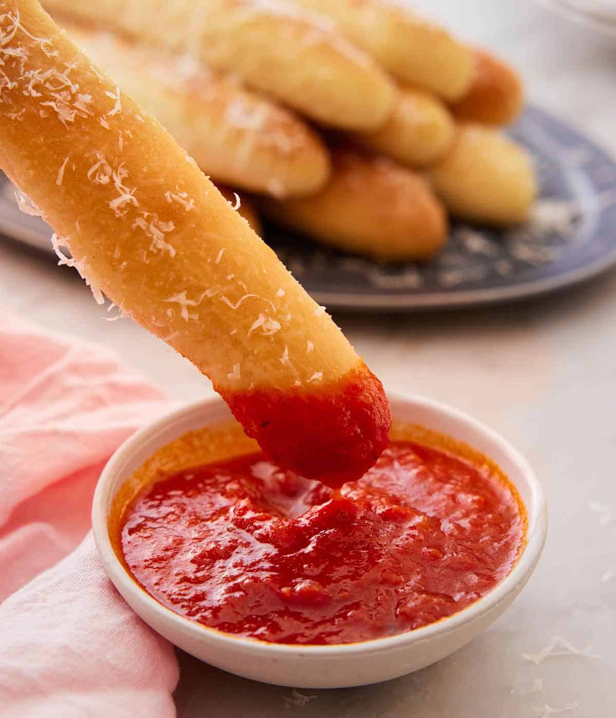 A breadstick dipped in a small bowl of red sauce with grated parmesan on the bread. More breadsticks in the background.