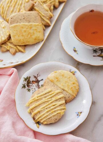A plate with two browned butter-earl grey cookies by a cup of tea and more cookies in the back.