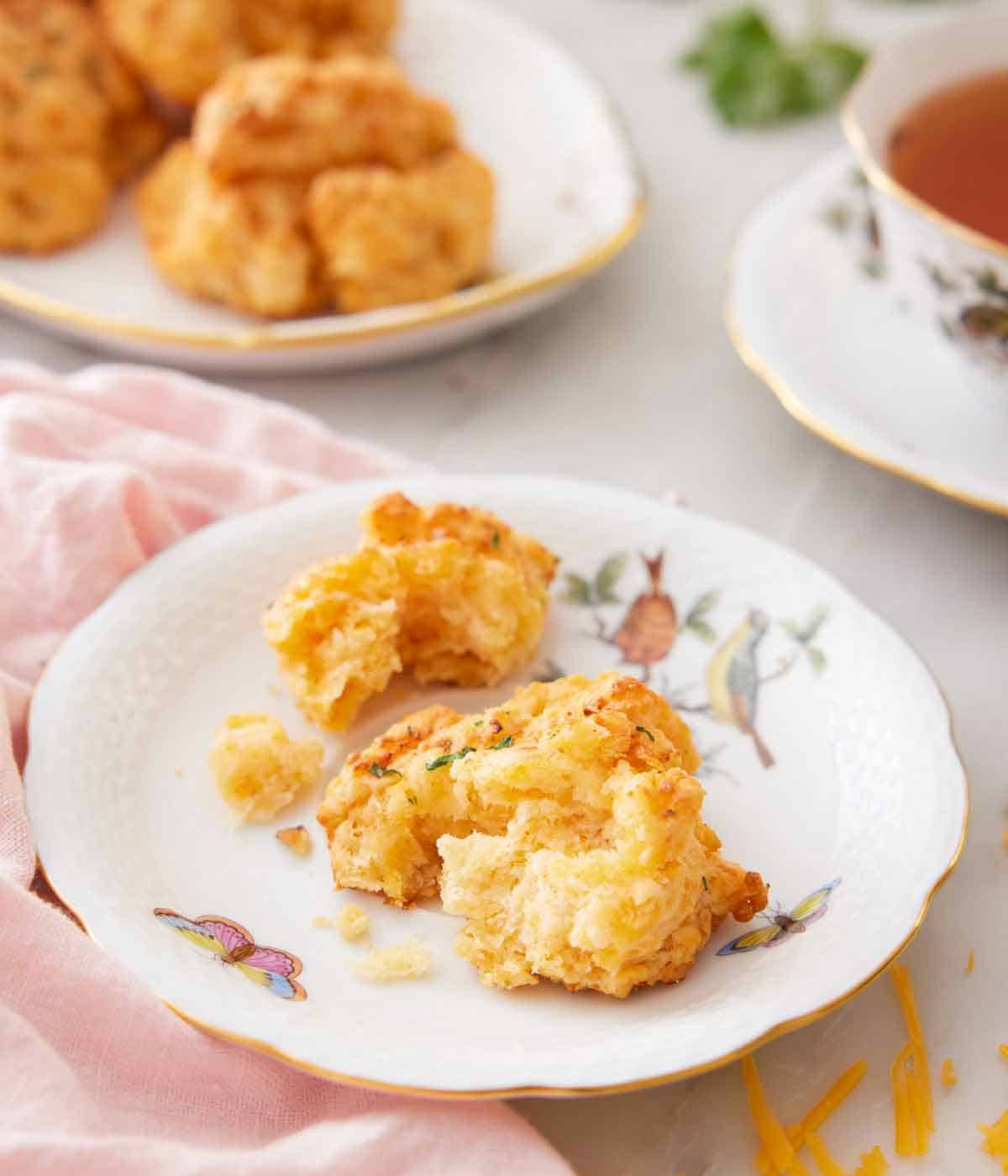 A plate with a cheddar biscuit torn apart with more biscuits and a cup of tea in the background.