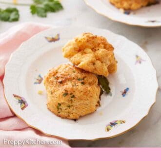 Pinterest graphic of a plate with two cheddar biscuits with another plate of two in the background along with some shredded cheese and parsley.