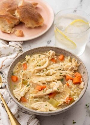 A bowl of chicken and rice soup with iced lemon water and bread in the background.