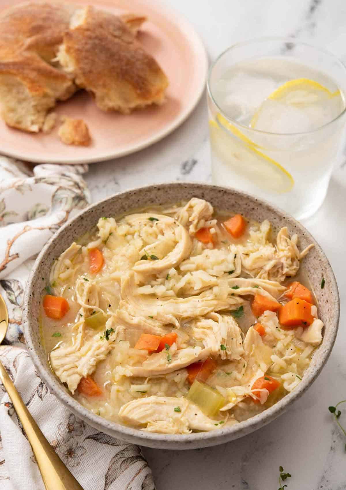 A bowl of chicken and rice soup with iced lemon water and bread in the background.