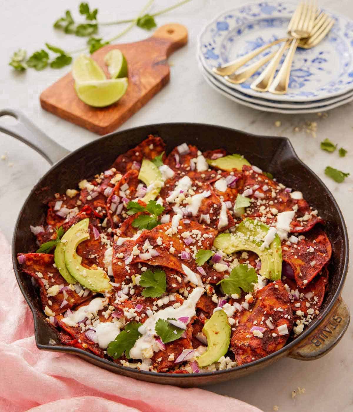 A cast iron skillet with chilaquiles with sliced avocado, creama, cotija, and cilantro garnished on top with cut limes, forks, and plates in the background.