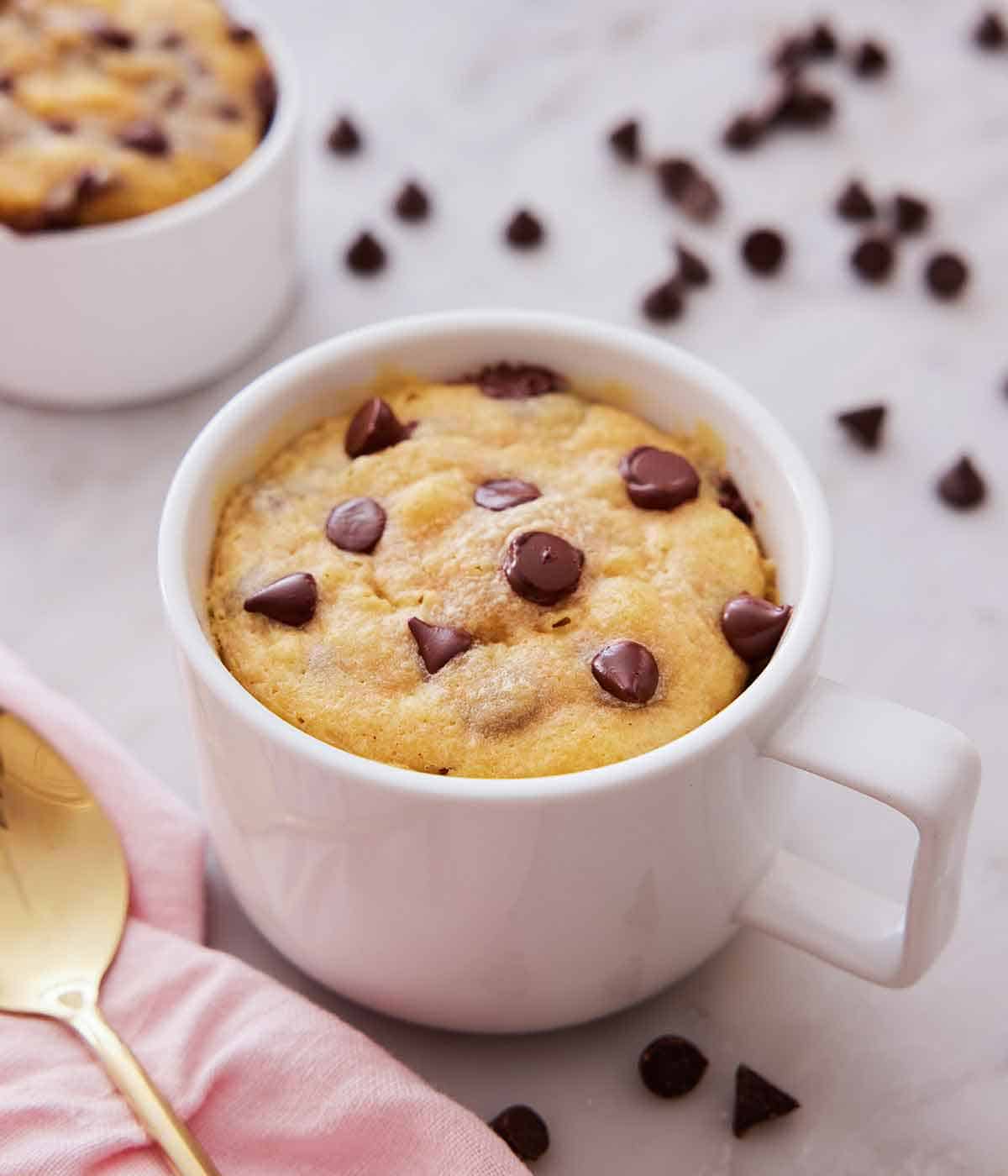 A cookie in a mug with chocolate chips scattered around with a spoon beside the mug.
