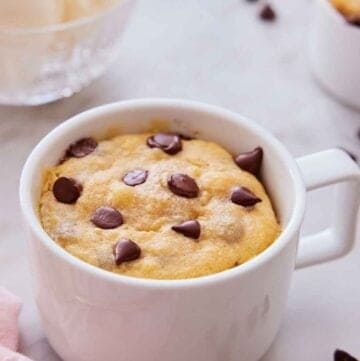 A cookie in a mug with chocolate chips scattered around.