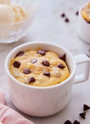 A cookie in a mug with chocolate chips scattered around.