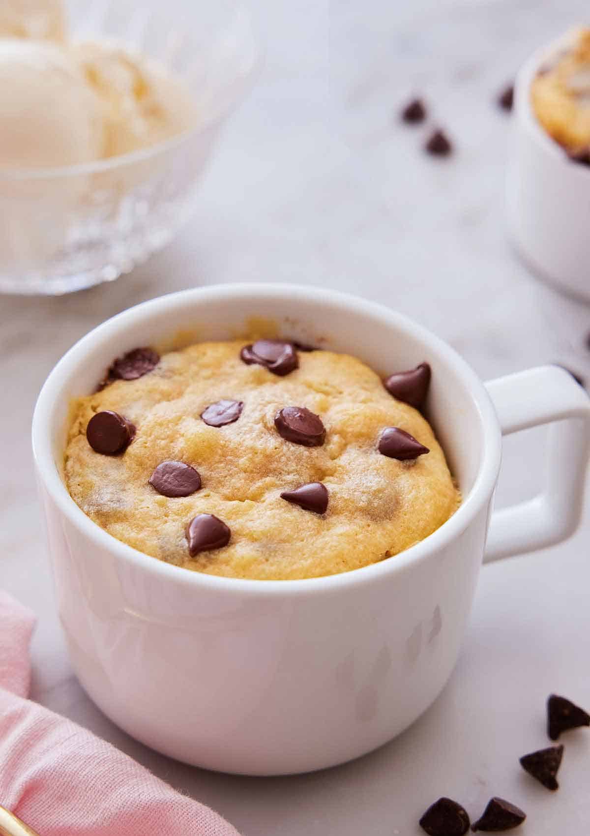 A cookie in a mug with chocolate chips scattered around.