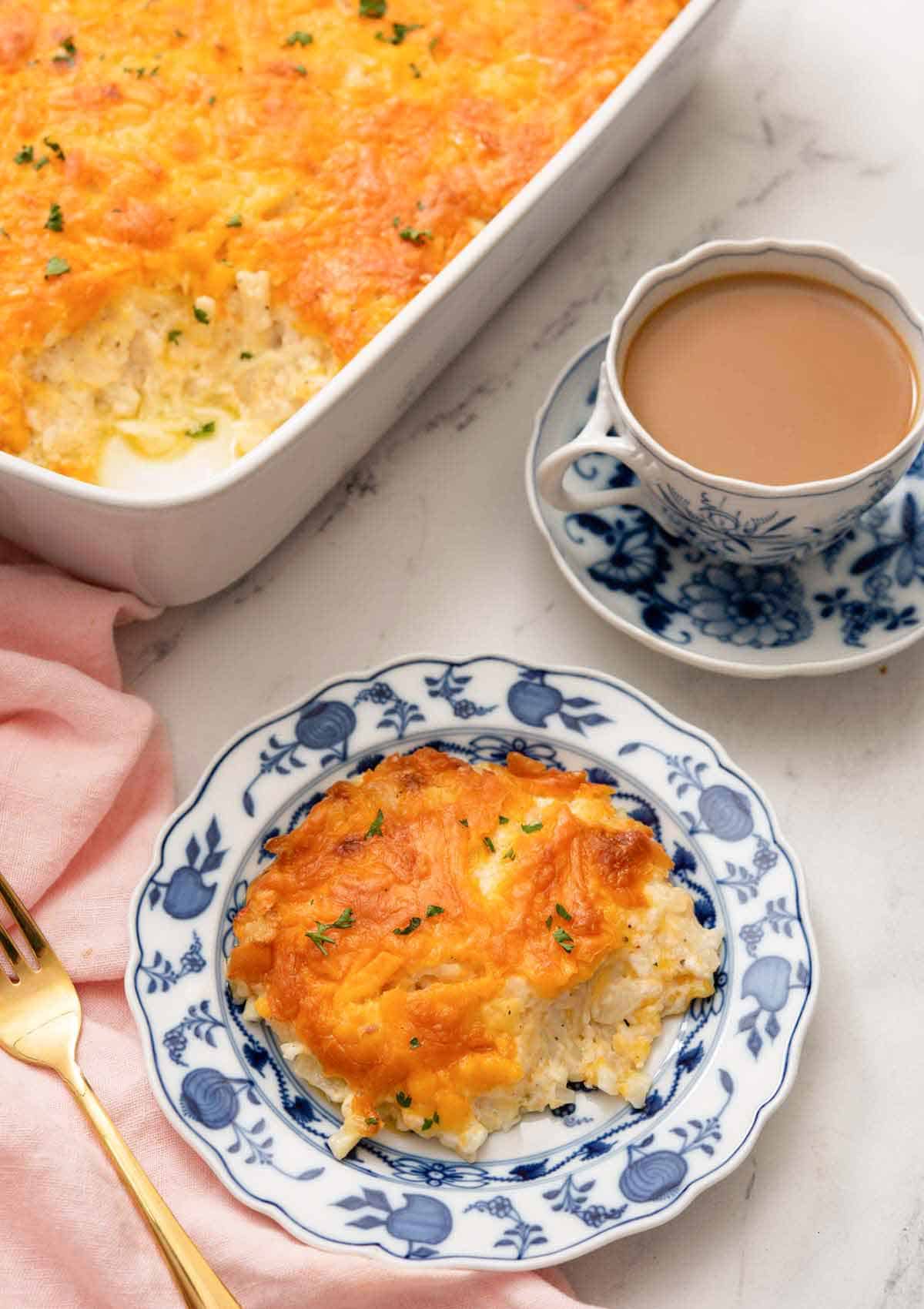 A plate with a serving of hashbrown casserole with a mug of coffee and the baking dish in the background.