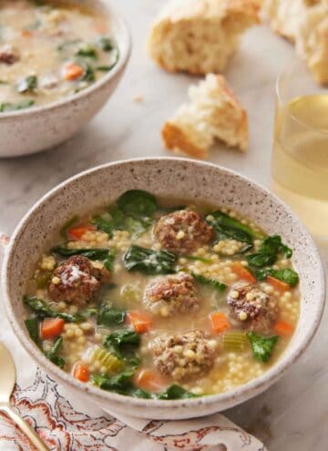 A bowl of Italian wedding soup with some torn bread and a glass of wine in the background.