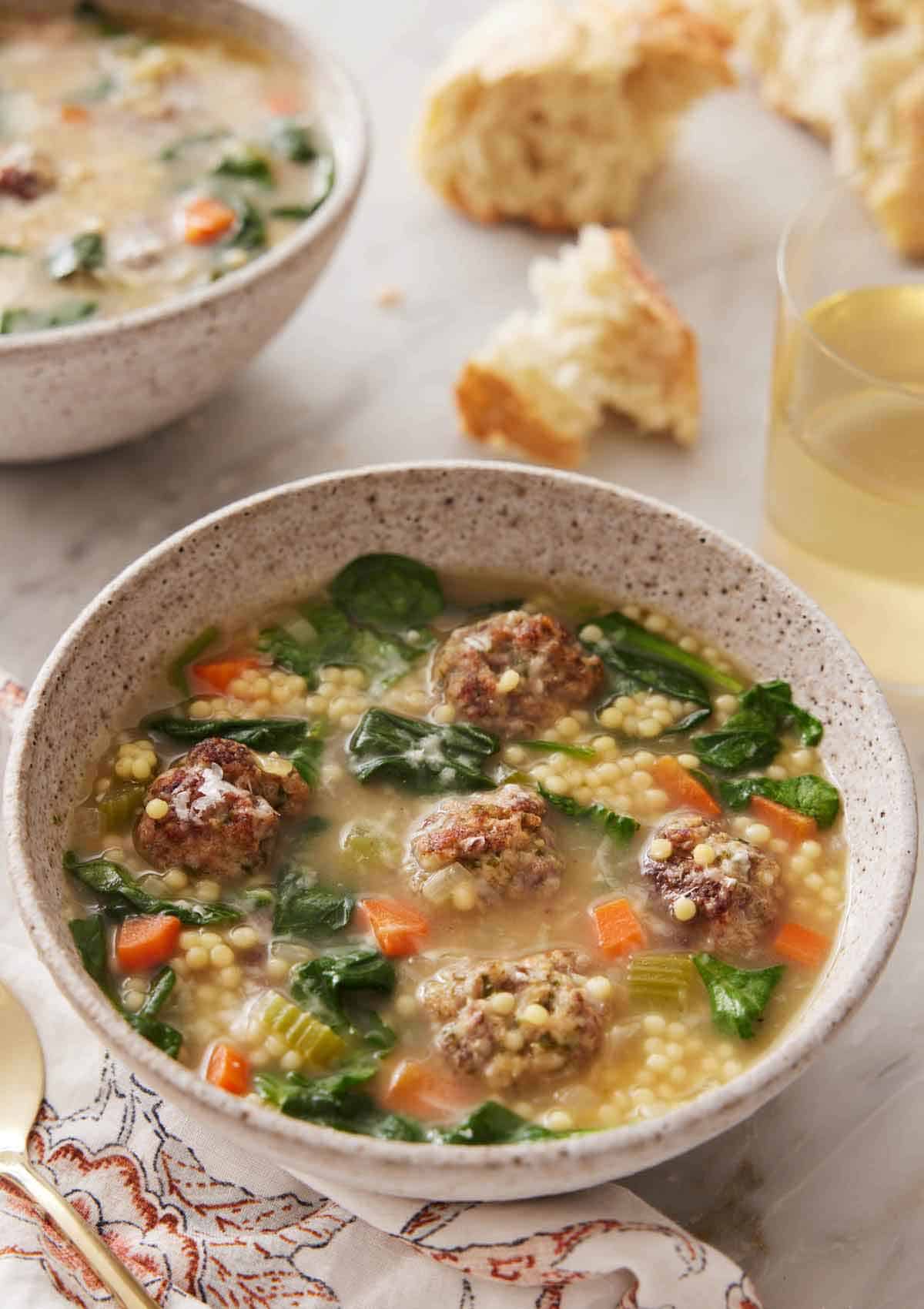 A bowl of Italian wedding soup with some torn bread and a glass of wine in the background.