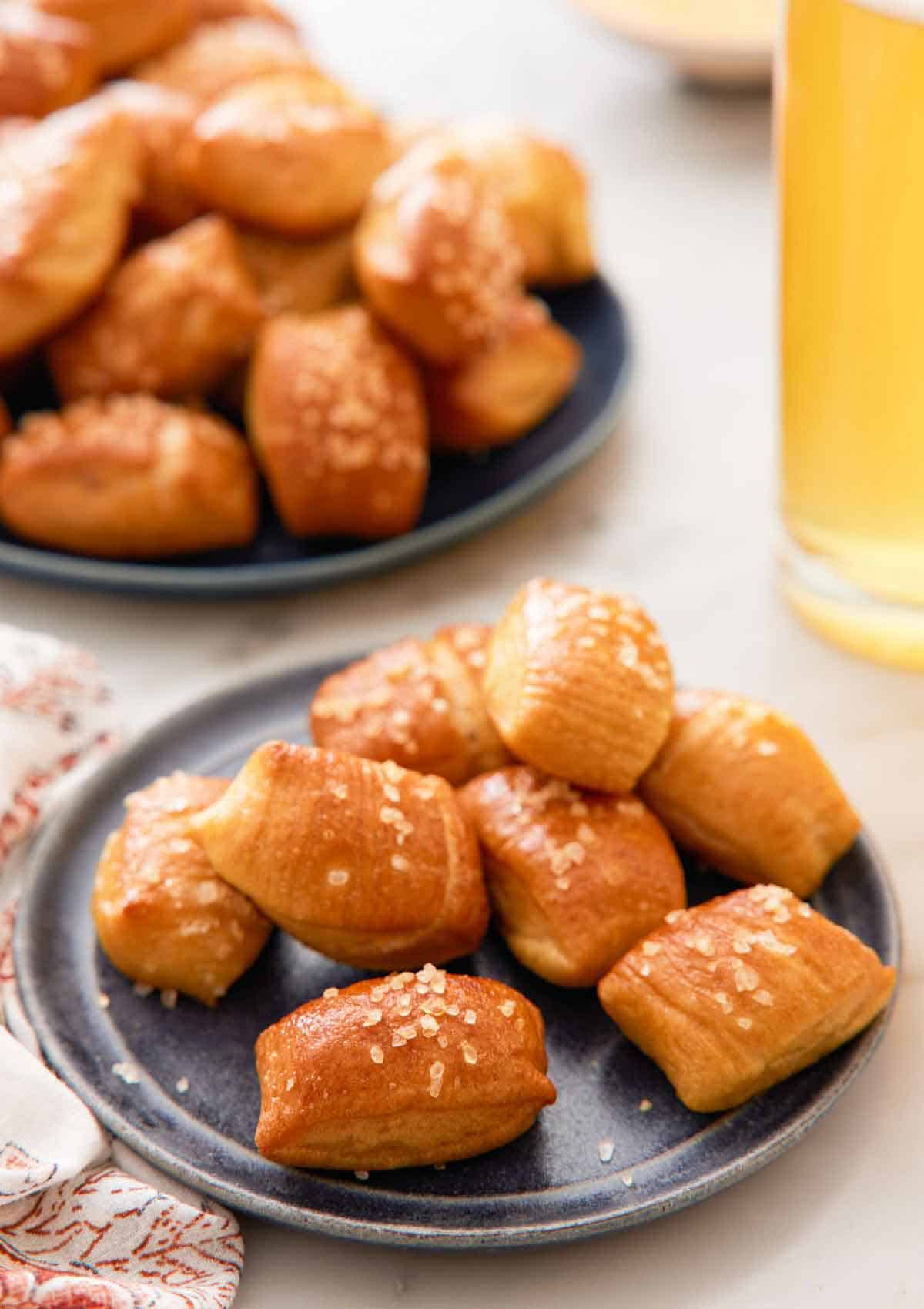 A plate of pretzel bites with a platter of more pretzel bites and a tall glass of beef in the back.