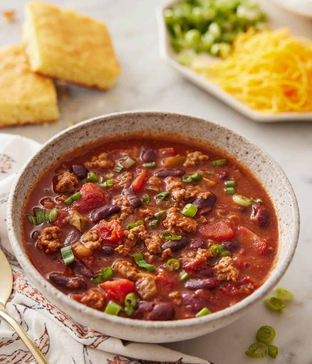 A bowl of slow cooker chili with toppings and cornbread in the background.