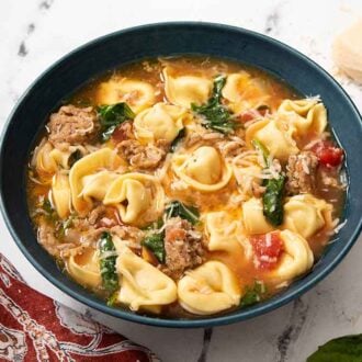 A blue bowl of tortellini soup with basil and parmesan in the background.