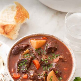Pinterest graphic of a bowl of beef stew with bread, water, and a pot in the background.