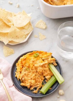A plate with buffalo chicken dip and two celery sticks with a plate of tortilla chips and a cup of water in the background.