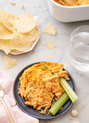 A plate with buffalo chicken dip and two celery sticks with a plate of tortilla chips and a cup of water in the background.