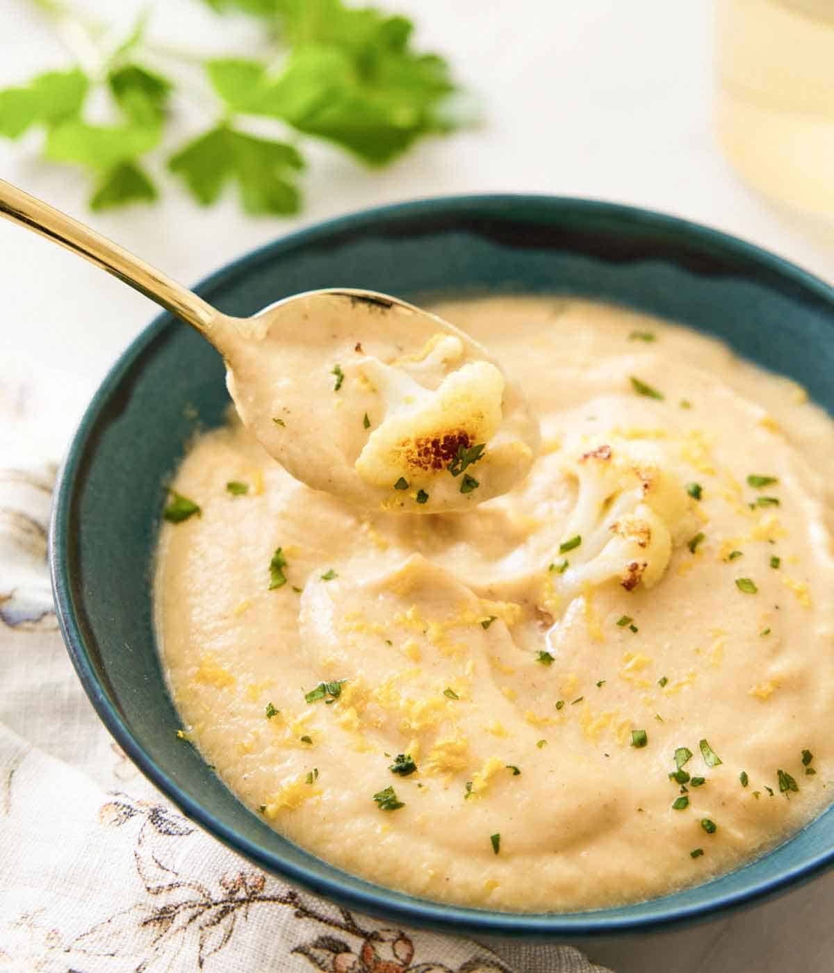A bowl of cauliflower soup garnished with parsley and lemon zest with a spoonful lifted out.