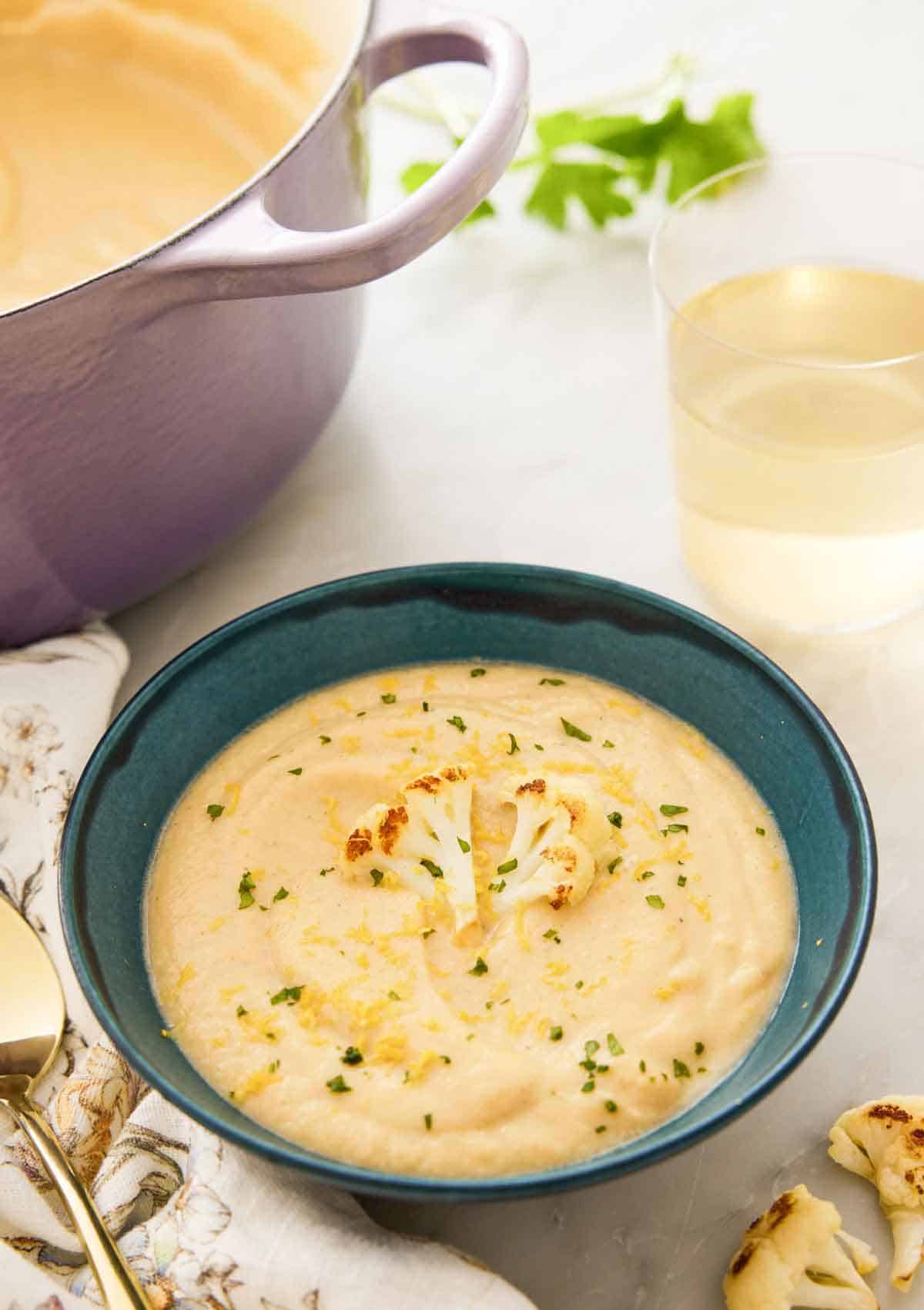 A bowl of cauliflower soup topped with a roasted cauliflower floret in front of a glass of wine and a dutch oven.