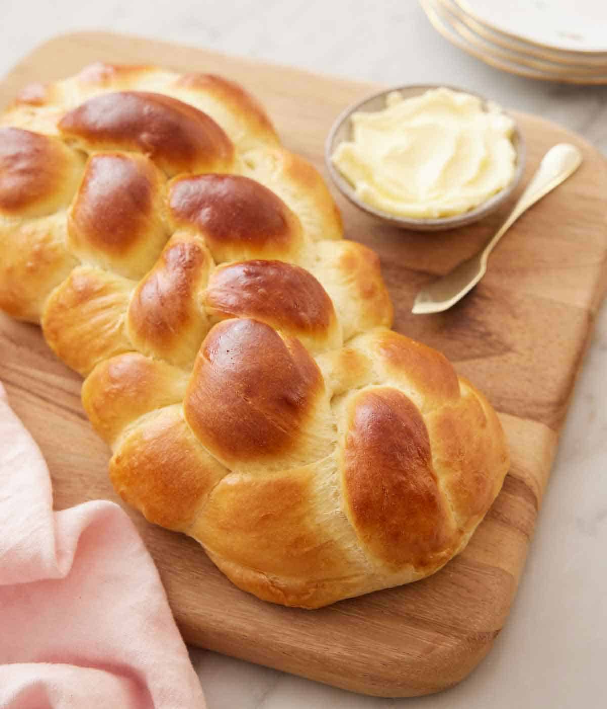 A loaf of challah on a wooden serving board with a bowl of butter with a spreader behind it.