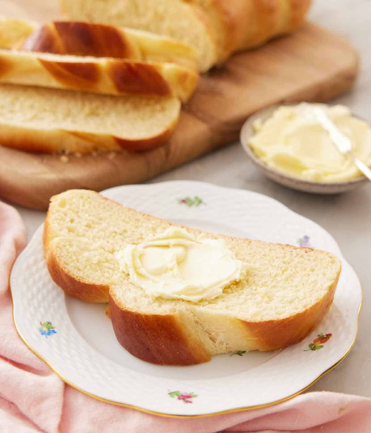 A plate with a slice of challah with a dollop of butter in the middle. Sliced challah on a serving board in the background with butter.