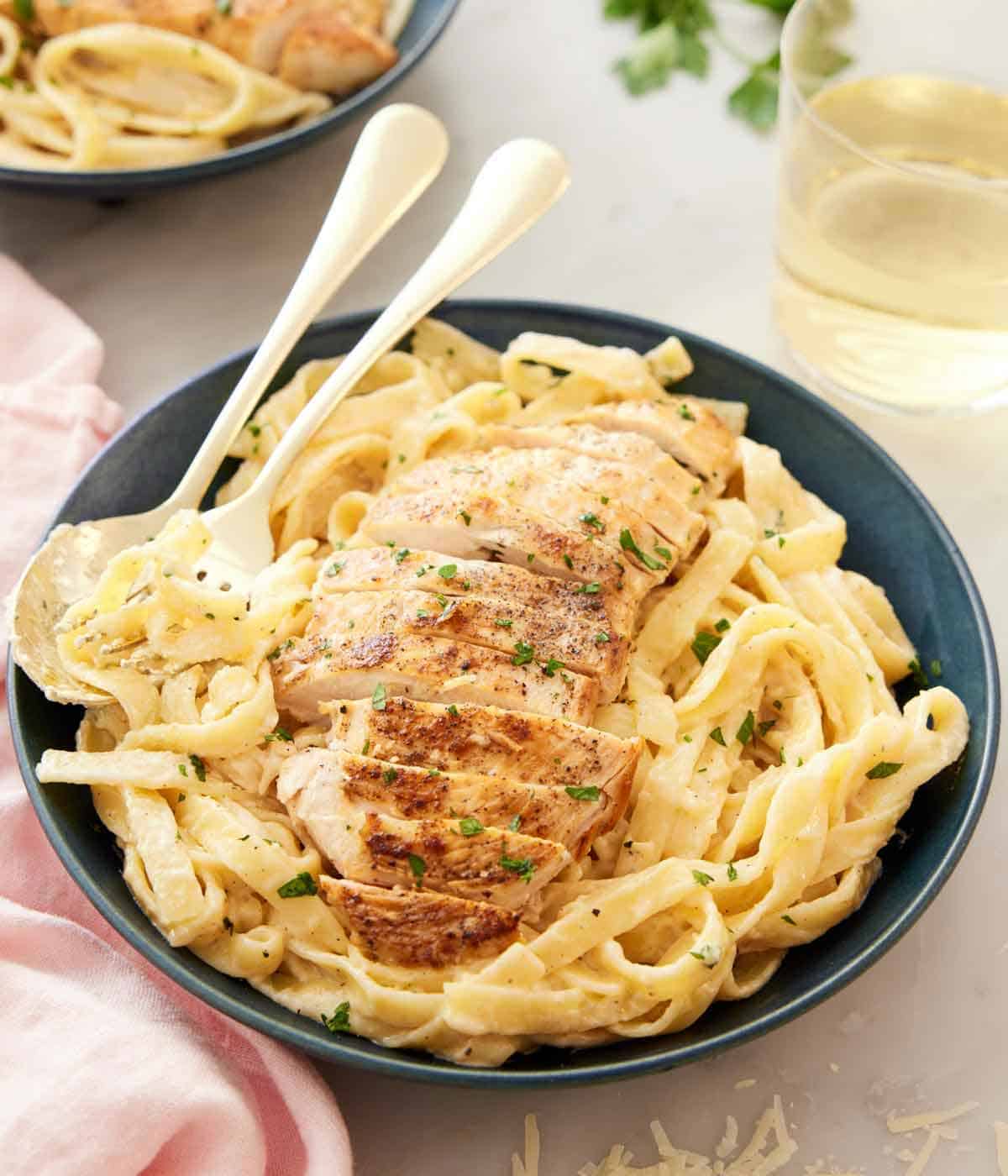 A bowl with a serving of chicken alfredo with the sliced chicken breast on top with a spoon and fork tucked in.