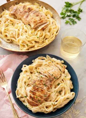 A plate of chicken alfredo pasta in a bowl in front of a glass of wine and a skillet with another serving of chicken alfredo.