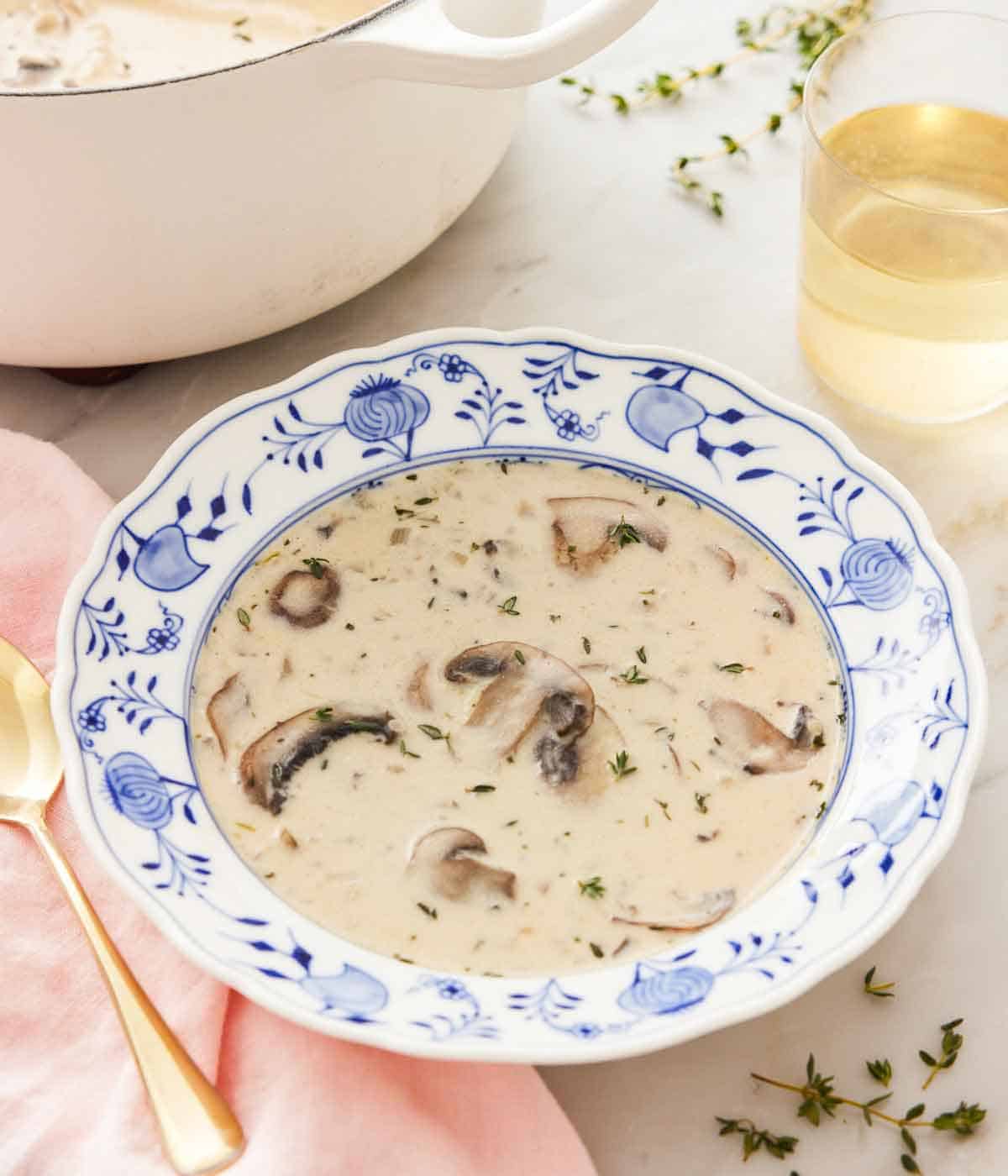 A bowl of cream of mushroom soup with a glass of wine and white pot in the background.