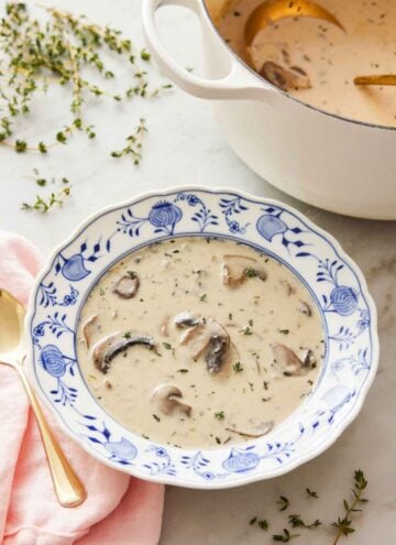 A bowl of cream of mushroom soup with fresh thyme on top and scattered around with a pot of soup in the background.
