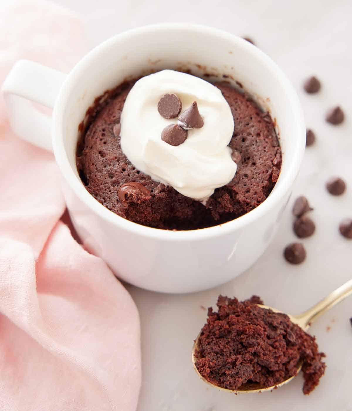 A mug cake with whipped cream and chocolate chips on top with a spoonful scooped out in front.