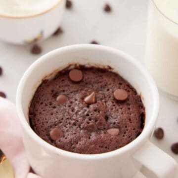 A mug cake with chocolate chips on top with more chocolate chips scattered in the back with a glass of milk.