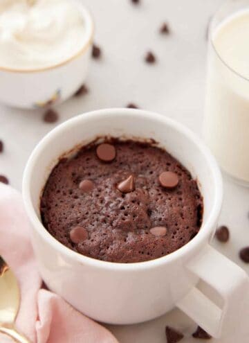 A mug cake with chocolate chips on top with more chocolate chips scattered in the back with a glass of milk.