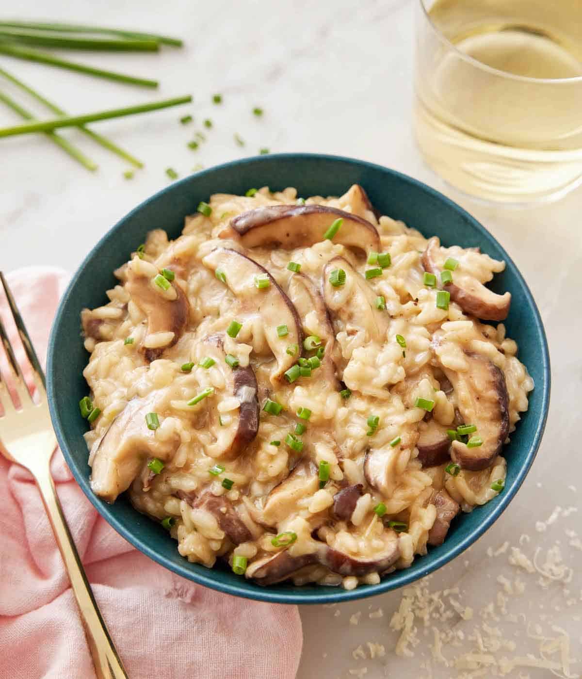 A bowl of mushroom risotto topped with chopped chives with a glass of wine, shaved parmesan, chives, and a fork surrounding it.