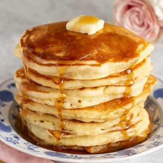 A plate with a stack of seven pancakes with syrup on drizzling down the pancakes and a slab of butter on top.