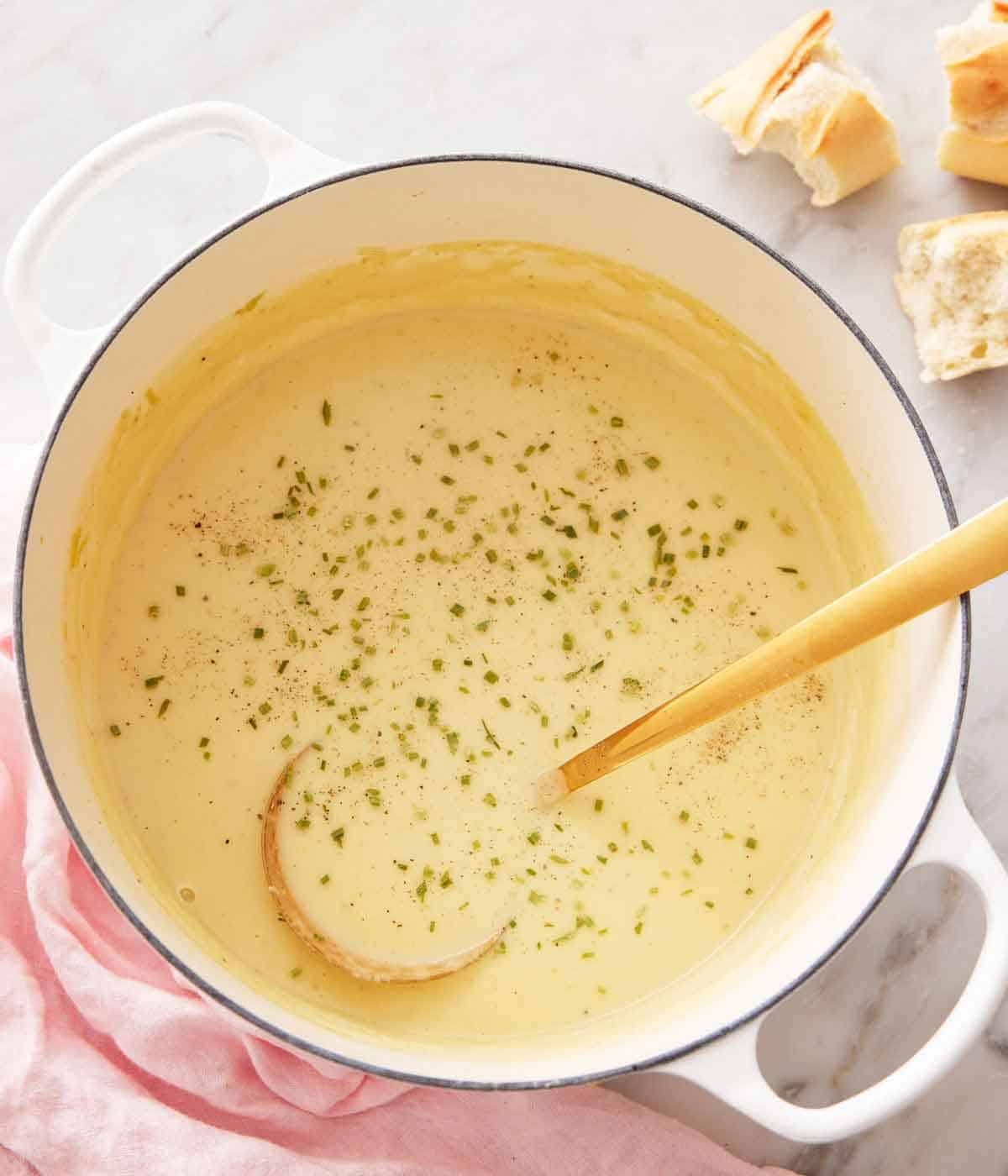 Overhead view of a white pot of potato leek soup with a ladle inside and some torn bread off to the side.