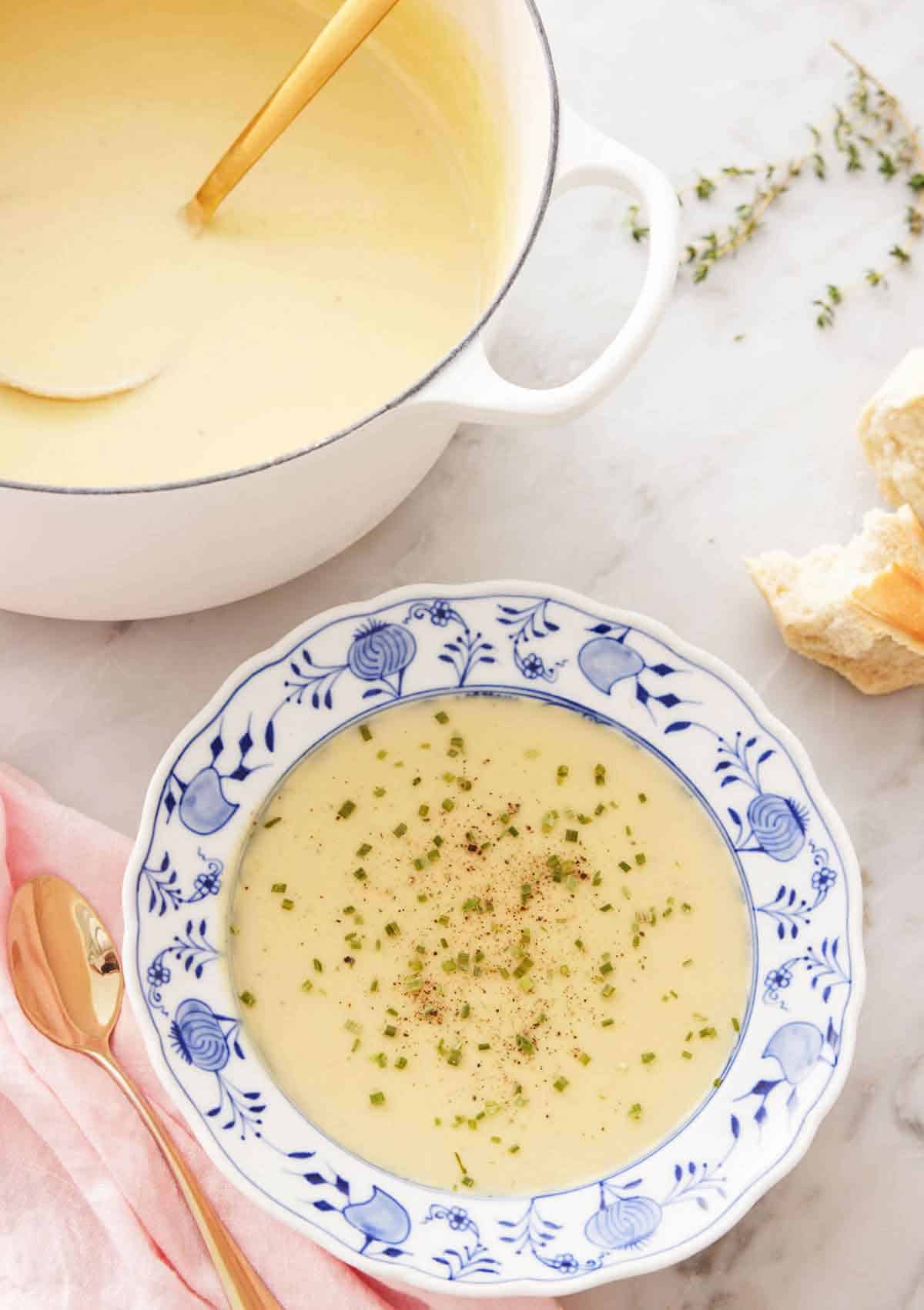 A bowl of potato leek soup with garnish with a large pot of soup with a ladle in the background.