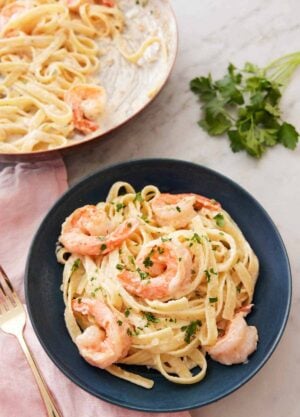 A plate of shrimp Alfredo with a bunch of parsley in the background along with a pan of the pasta.