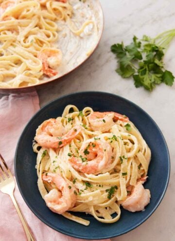 A plate of shrimp Alfredo with a bunch of parsley in the background along with a pan of the pasta.