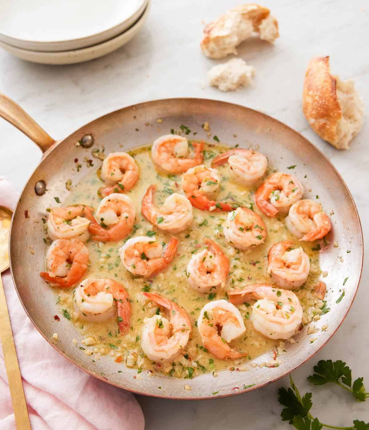 A skillet holding shrimp scampi with some torn bread in the background with some plates.