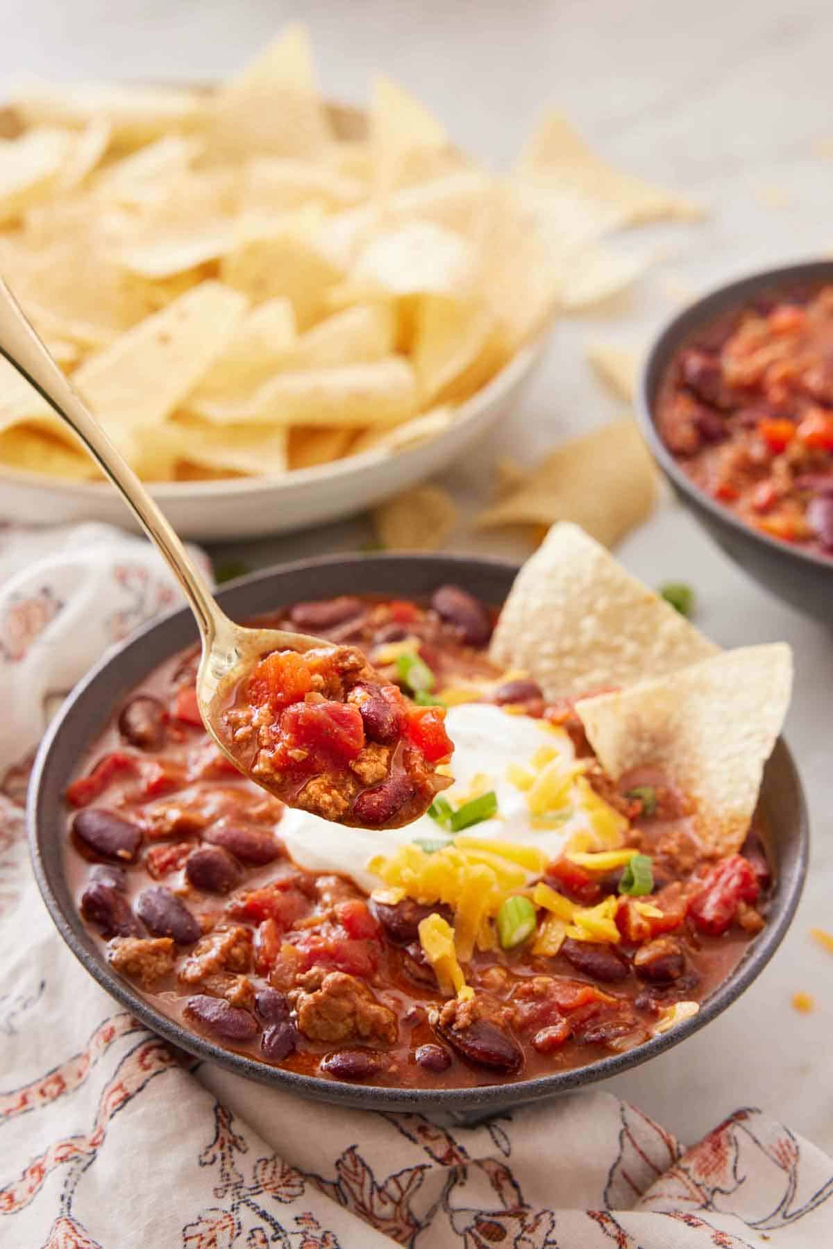 A spoonful of turkey chili lifted from a bowl. A bowl of chips in the background.