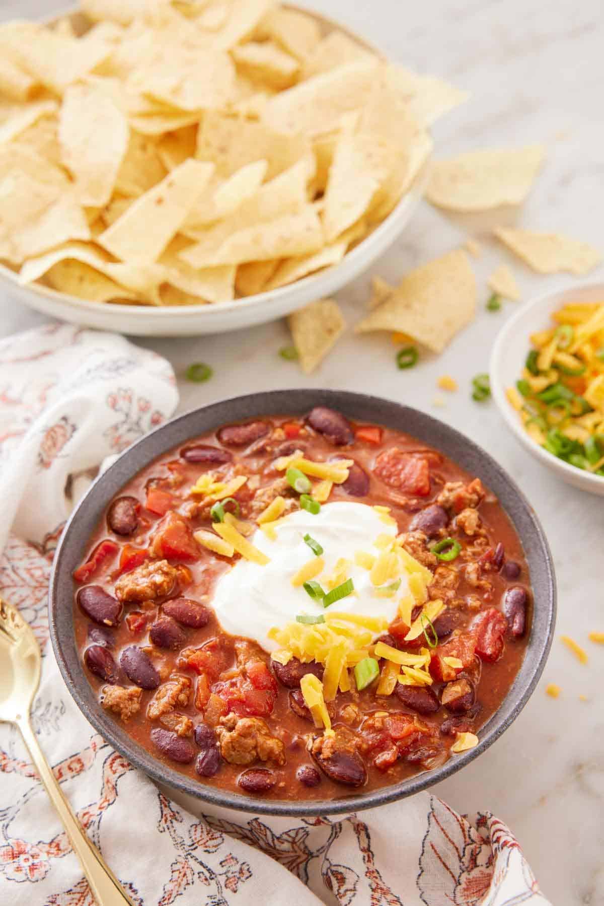 A bowl of turkey chili topped with sour cream and cheese with a bowl of chips and bowl of toppings in the back.