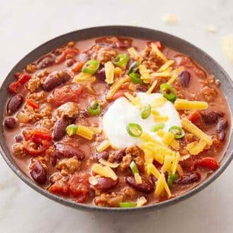 A bowl of turkey chili topped with a dollop of sour cream, shredded cheese, and green onions. Chips scattered in the back.
