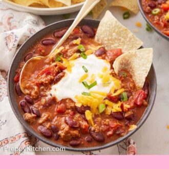 Pinterest graphic of a bowl of turkey chili topped with sour cream, cheese, green onions, and two chips with a spoon tucked in.