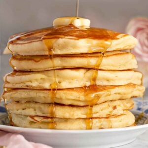 A tall stack of pancakes topped with a pat of butter with syrup being poured on top.