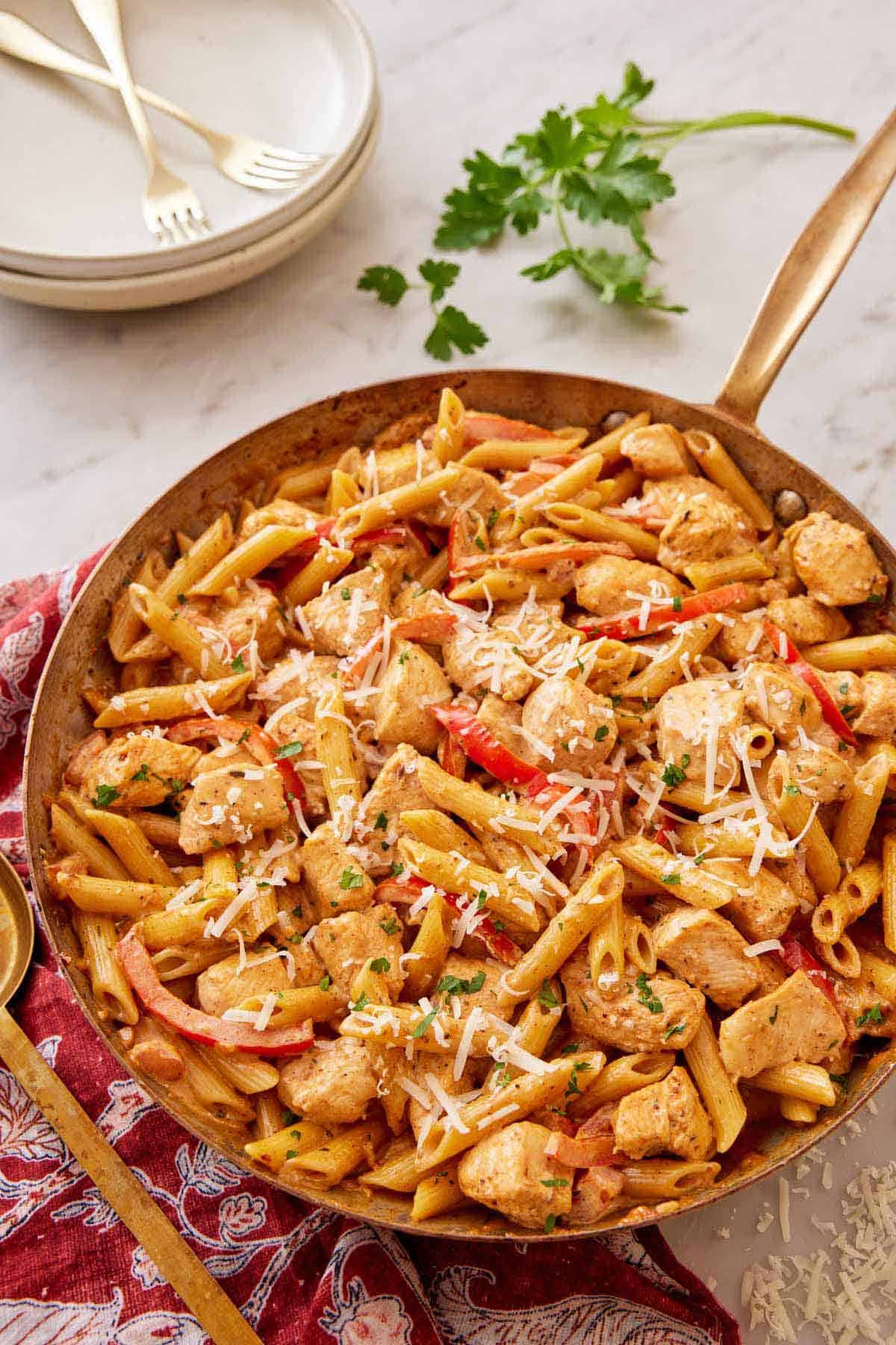 A skillet of Cajun chicken pasta with freshly shredded parmesan on top. Some parsley and a plate and fork in the back.