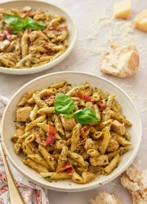 A plate of chicken pesto pasta with basil and sun-dried tomatoes with some bread, shredded parmesan and another plate of pasta in the background.