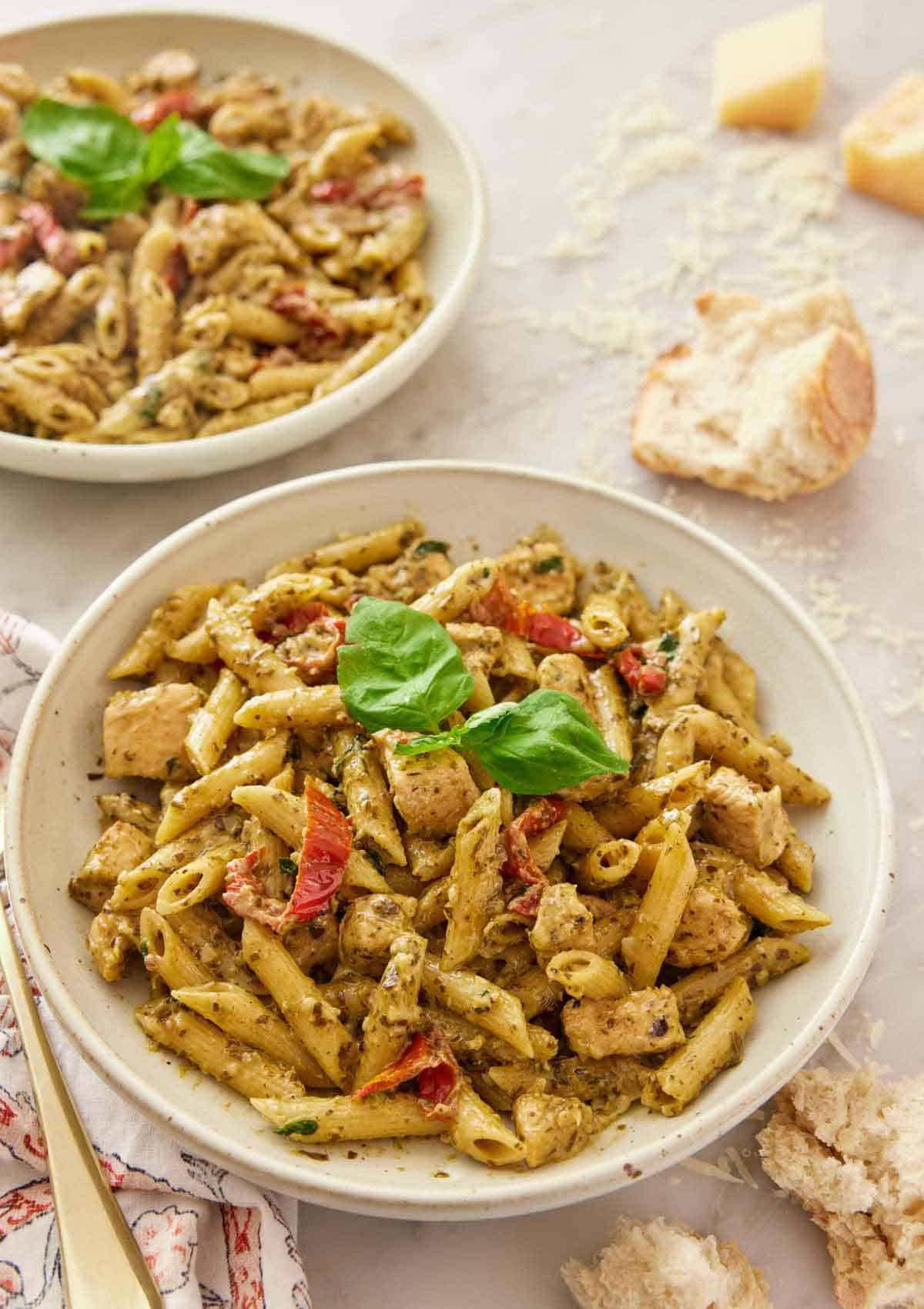 A plate of chicken pesto pasta with basil and sun-dried tomatoes with some bread, shredded parmesan and another plate of pasta in the background.