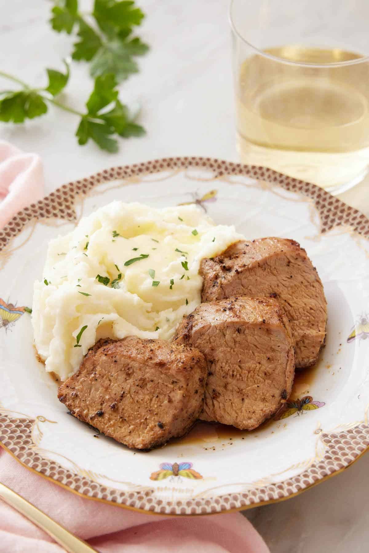 A plate with three pieces of pork tenderloin with mashed potatoes on the side with a glass of wine in the background.