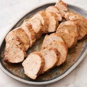 A grey platter with sliced pork tenderloin with some juices on the platter.