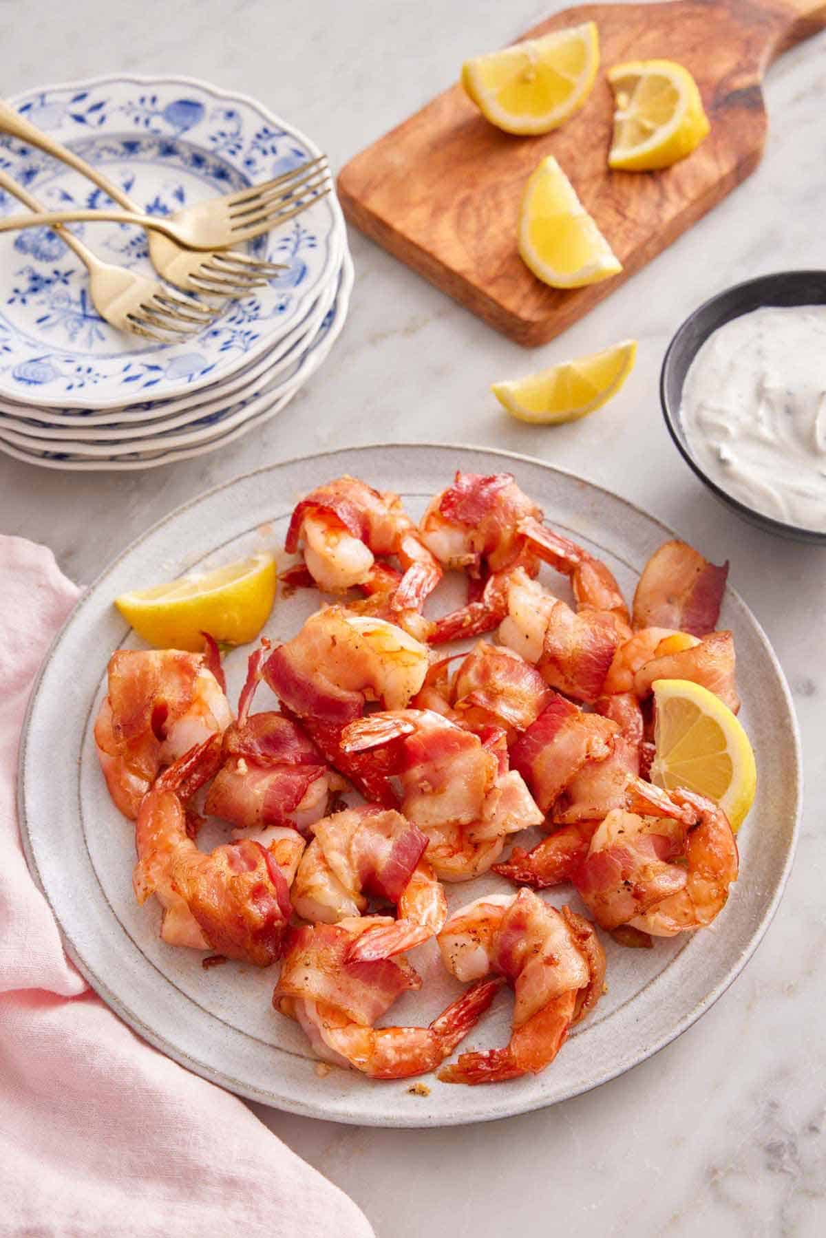 A plate of bacon wrapped shrimp with two lemon wedges with dip, more lemons, plates, and forks in the back.