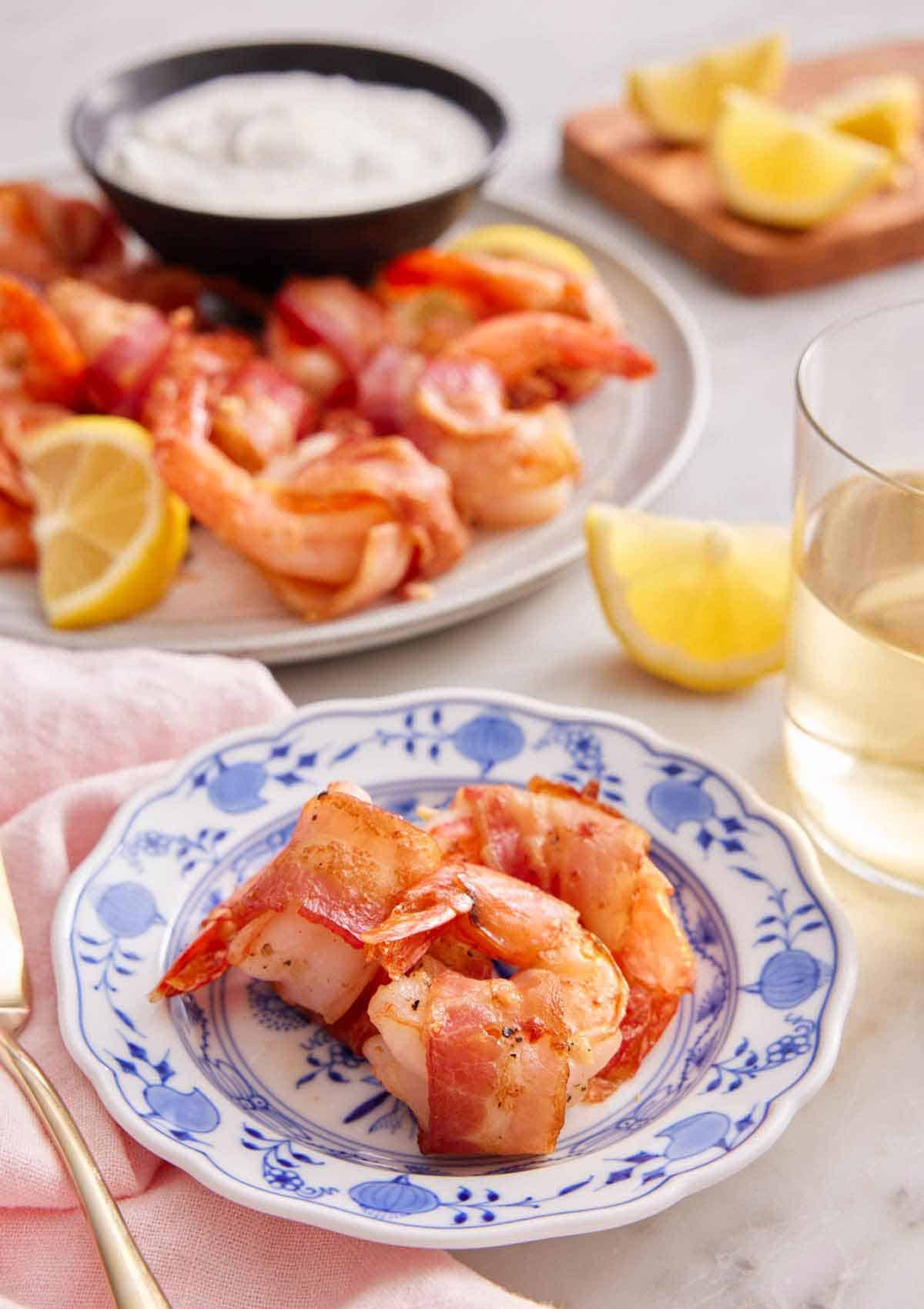 A plate with three pieces of bacon wrapped shrimp with more in the back with dip, lemons, and a glass of wine.